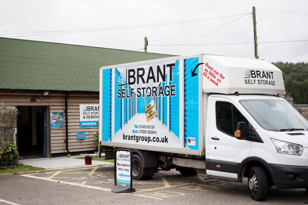 A white Brant Self Storage van is parked outside a wooden storage facility. The van displays the company logo and website. A sign in front reads Reception this way, with the facility building in the background.