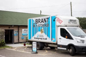 A white Brant Self Storage van is parked outside a wooden storage facility. The van displays the company logo and website. A sign in front reads Reception this way, with the facility building in the background.