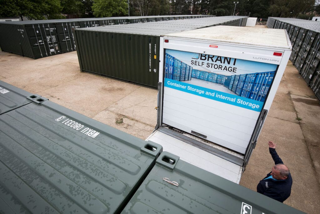 A man stands near a shipping container at a self-storage facility with rows of green containers and a white storage truck labeled Brant Self Storage. Trees are visible in the background.